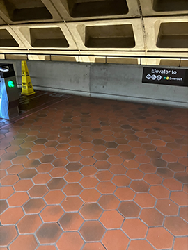 A picture of the entrance of the guided pathway next to the faregates. To the left there is a yellow wet floor cone next to a faregate and to the right in the background there is a sign pointing to the elevator on the wall. The wall in the background has a railing on top of it.